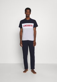 Námornícky modré a šedé tričko značky Lacoste s červeným logom, v kombinácii s námornícky modrými nohavicami. Uvoľnený strih, bavlnený materiál, jednoduchý dizajn.