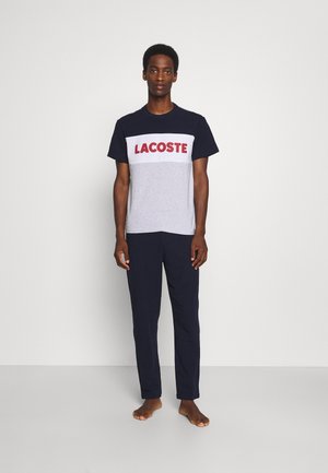 Tricou Lacoste de culoare bleu marin și gri, cu logo roșu, asortat cu pantaloni bleu marin. Croi lejer, material din bumbac, design simplu.