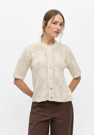 Femme portant un cardigan en tricot crème à manches courtes avec des boutons, associé à un pantalon marron foncé sur un fond blanc uni.