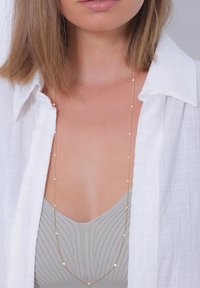 Guld-tone halskæde med forlænget kæde, prydet med små, vekslende klare og gyldne perler. Bæret over en grå top og en hvid skjorte.