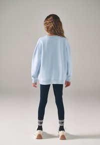 Šviesiai mėlynas per didelis džemperis su nuleistais pečiais, derinamas su tamsiai mėlynomis tamprėmis ir baltais sportbačiais su tekstūruotais padais ir juodais akcentais.