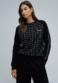 Sweatshirt noir avec un motif de grille texturé, présentant des manches noires contrastantes et des accents métalliques sur le côté.