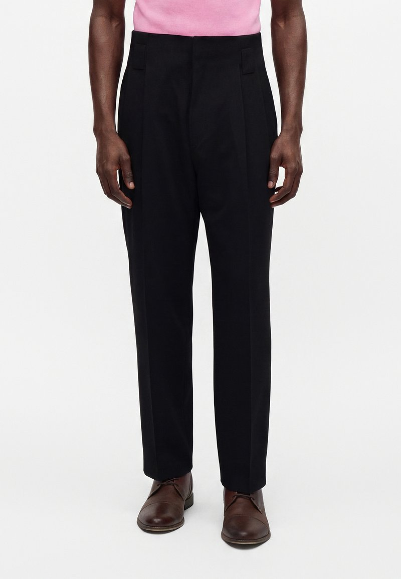 Homme portant un pantalon noir sur mesure, des chaussures en cuir marron et une chemise rose, debout devant un fond clair uni.