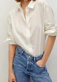 Camisa blanca de botones hecha de un tejido suave con mangas arremangadas, combinada con unos vaqueros de mezclilla azul de cintura alta con un corte clásico y costuras.