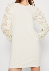 Roomkleurige jurk met een ronde hals en lange doorschijnende mouwen versierd met stoffen bloemapplicaties. Zachte, gladde textuur.