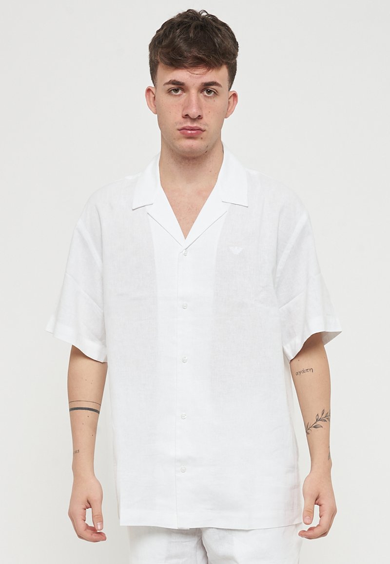 Camicia bianca in lino a maniche corte con colletto, dotata di bottoni frontali e di un piccolo logo sul petto. Tessuto liscio e vestibilità comoda.