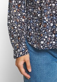 Blouse bleu marine à fleurs avec manches longues, ornée de fleurs blanches, bleues et rouille. Manches à revers et coupe décontractée, portée avec un jean bleu.