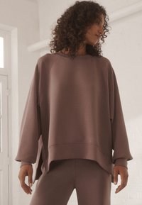 Felpa oversize color malva tenue, con maniche raglan, scollo ampio e tessuto testurizzato con orlo leggermente curvo.