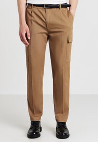 Pantaloni cargo marroni realizzati in tessuto leggero. Presentano tasche laterali, una vestibilità slim e un davanti piatto con una cintura nera. Le scarpe nere completano il look.