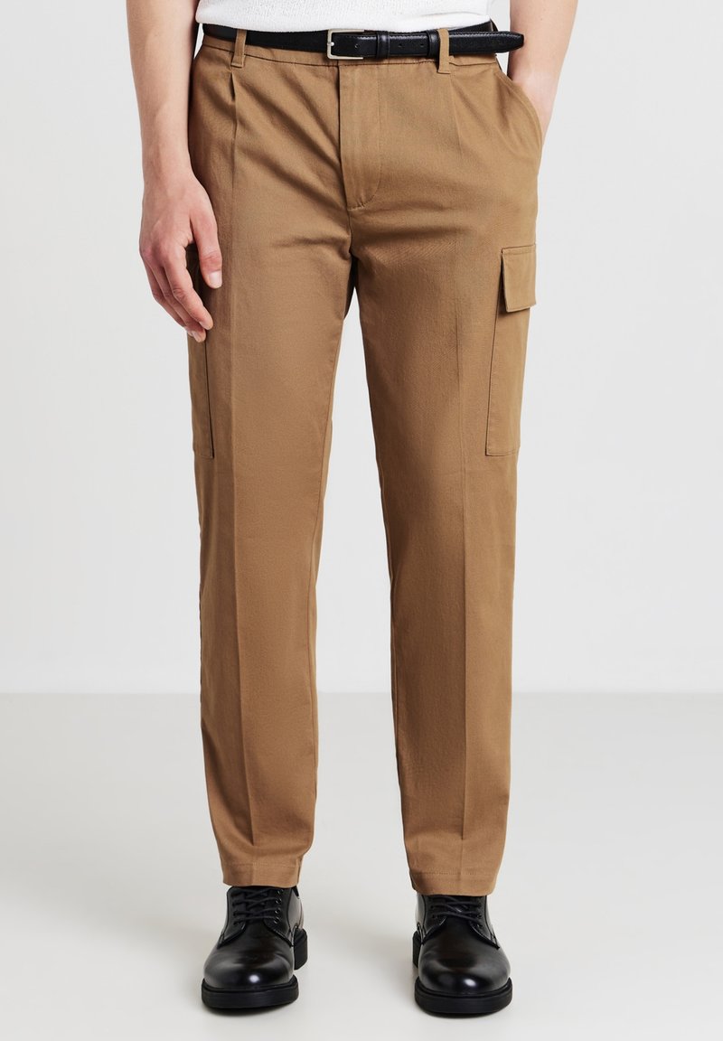 Pantaloni cargo marroni realizzati in tessuto leggero. Presentano tasche laterali, una vestibilità slim e un davanti piatto con una cintura nera. Le scarpe nere completano il look.