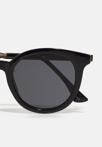 Ochelari de soare rotunzi, negri, cu rame groase și glosate și lentile închise la culoare. Prezintă accesorii din metal auriu pe brațe și un design elegant.