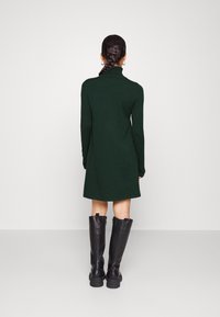 Robe en maille verte avec col montant et manches longues, associée à de grandes bottes en cuir noir. Texture lisse, longueur aux genoux.