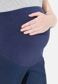 Graviditetströja i grått med vita prickar och en marinblå stretchpanel, med mjukt tyg och en figurnära design. Handen vilar på magen.