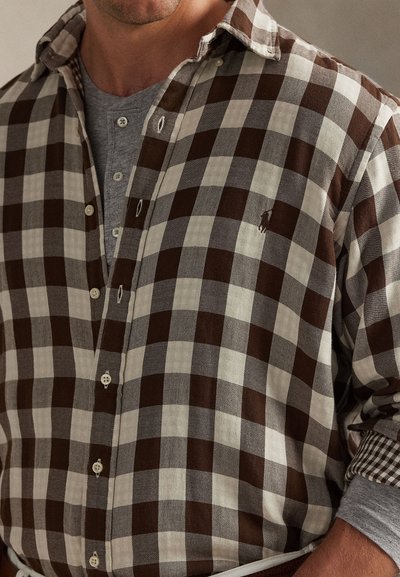 Homem a usar uma camisa de botão às quadrículas castanha e branca sobre uma camisa henley cinzenta, com as mangas enroladas de forma descontraída.
