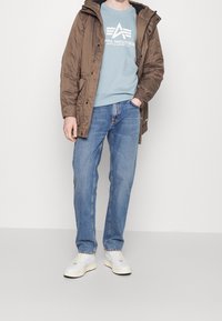 Brun isolerad jacka, ljusblå sweatshirt med vitt tryck, blå raka jeans och vita sneakers på en neutral bakgrund.