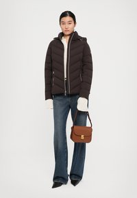 Kvinne iført en brun boblejakke, hvit genser, blå jeans, svarte spisse sko, som holder en brun skinnveske i stroppen.