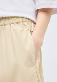 Δεξί χέρι τοποθετημένο στην πλαϊνή τσέπη μπεζ παντελονιών με ελαστική μέση, φορεμένων με λευκό μπλουζάκι κοντομάνικο.