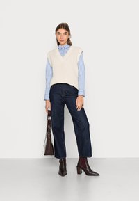 Gilet lavorato a maglia beige chiaro con scollatura a V, camicia a righe blu e bianche, jeans a vita alta blu scuro e stivaletti alla caviglia borgogna. Borsa marrone.