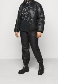 Czarna kurtka puchowa o błyszczącej powierzchni, zestawiona z czarnym t-shirtem z grafiką i czarnymi skórzanymi spodniami. Całość dopełniają masywne czarne buty.