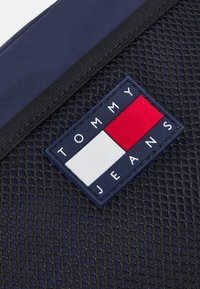 Detalj av marinblå tygväska med en gummimärke som har röda, vita och blå rektanglar, och "TOMMY JEANS" präglat i vita bokstäver.