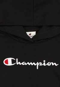 Sudadera negra de algodón, con un logo "Champion" bordado en blanco y un emblema rojo. Textura suave, diseño de capucha estándar.