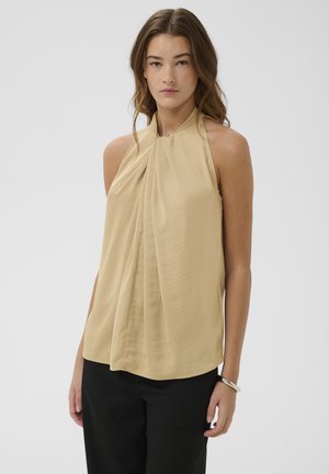 Junge Frau mit langen braunen Haaren, die ein beiges ärmelloses plissiertes Neckholder-Top und schwarze Hosen vor einem einfarbigen weißen Hintergrund trägt.