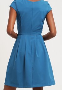 Eine Frau trägt ein ärmelloses, knielanges blaues Kleid, von hinten gezeigt, mit den Händen auf der Taille.