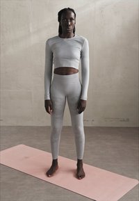 Crop top cu mânecă lungă de culoare gri deschis, cu panouri texturate, alături de colanți cu talie înaltă asortați, așezați pe un covoraș de yoga roz, având un design ajustat.