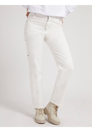 Person iført hvide, lige bukser og beige snørestøvler til ankler, stående med hænderne i lommerne mod en ensfarvet baggrund.