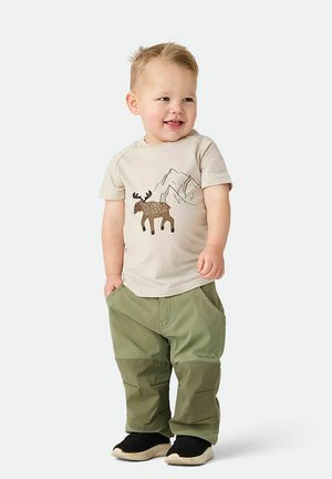 Småbarn med kort blondt hår iført beige t-shirt med elg- og bjergprint, grønne bukser og sorte slip-on sko, stående og smilende.