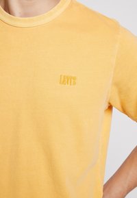 Žluté tričko s kulatým výstřihem a malým vyšitým logem Levi's na levé hrudi, které má na sobě osoba s rukou na kyčli.