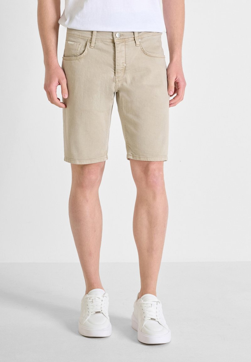 Pantalones cortos de mezclilla beige con cinco bolsillos, un botón en la parte delantera y un corte recto. Combinados con zapatillas blancas con cordones, que presentan una textura suave.