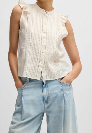 Crèmekleurige blouse met ruches aan de mouwen, voorzien van verticale plooien en knoopsluiting. Gecombineerd met lichtblauwe jeans met wijd uitlopende pijpen.