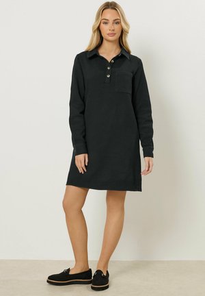 Kvinde står iført en sort skjortekjole med lange ærmer, forreste knapper og en brystlomme, kombineret med sorte slip-on sko.