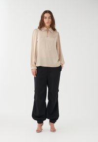 Beige højhalset bluse med en nøglehuldetalje, parret med løse sorte bukser med samlede cuffs og strop-heller.
