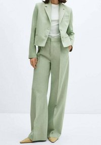 Costume vert clair avec un blazer ajusté et un pantalon à jambes larges, fabriqué en tissu texturé, associé à une chemise blanche unie et des chaussures pointues jaunes.