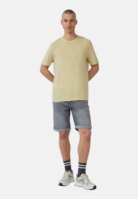 Hellgelbes T-Shirt mit kurzen Ärmeln, kombiniert mit grau-denim Shorts mit umgeschlagenen Säumen und weißen Sneakers, die mit schwarz-grau gestreiften Socken getragen werden.