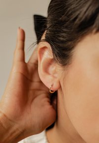 Boucles d'oreilles en or avec une surface texturée et de petites pierres multicolores, portées à l'oreille, avec une main touchant doucement l'oreille.