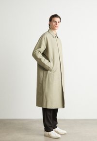 Licht olijfgroene trenchcoat met een recht gesneden model, zijzakken en een kraag, gecombineerd met zwarte broek en witte sneakers. Glad textuur.