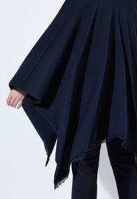 Dunkelblauer Poncho aus weichem Stoff, mit fließendem Design, unregelmäßigem Saum und fransigen Kanten. Eine Hand teilweise sichtbar.