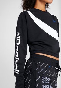 Felpa nera a taglio corto con una striscia diagonale bianca, con dettagli del logo blu e testo stampato sulle maniche. Abbinata a pantaloni neri.