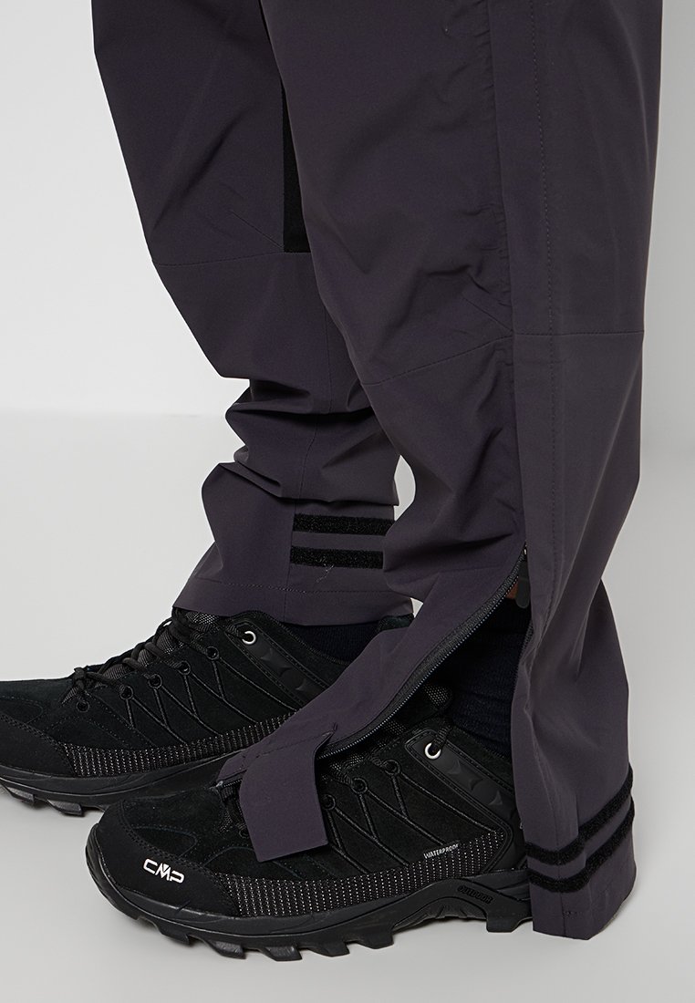 Zwarte wandelschoenen met gestructureerde accenten en veters, gecombineerd met paarse waterdichte broek met ritsen en gestreepte details.