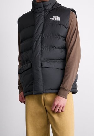 Mann trägt eine schwarze North Face gesteppte Weste über einem braunen Langarmshirt und gelben Hosen, steht vor einem schlichten Hintergrund.