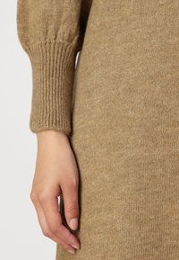 Stickad tröja i beige ullblandning med ribbad, uppvikt ärm och mjuk textur. Fokuserad bild av en hand som vilar mot plagget.