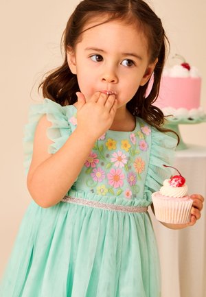 Junges Mädchen in mintgrünem Blumenkleid isst einen Cupcake mit weißem Zuckerguss und einer Kirsche darauf; im Hintergrund steht eine passende Torte.