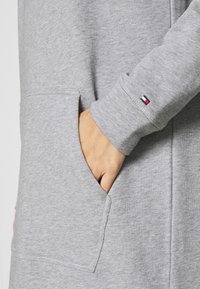 Tejido de sudadera gris con un bolsillo frontal. Presenta puños de manga acanalados y un pequeño logo con franjas rojas, blancas y azules en el puño.
