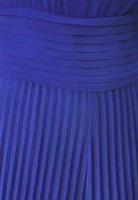 Złożona niebieska tkanina sukienki o teksturowanej powierzchni. Posiada szeroki pasek w talii i jednorodne pionowe fałdy na całej długości odzieży.