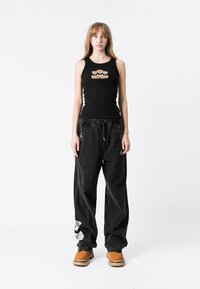 Top sin mangas negro con bordado floral y pantalones holgados negros con un parche de oso de peluche, combinados con zuecos marrones.
