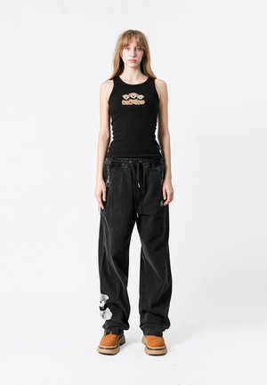 Top sin mangas negro con bordado floral y pantalones holgados negros con un parche de oso de peluche, combinados con zuecos marrones.