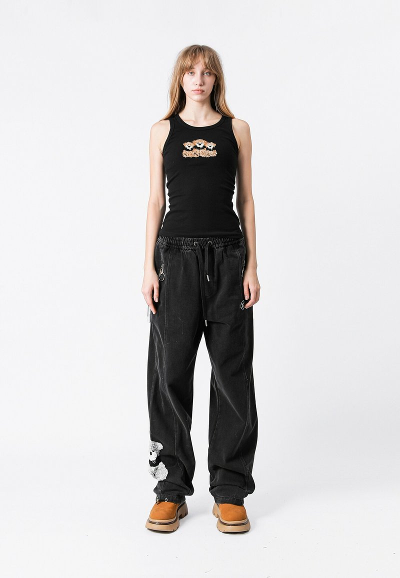 Top sin mangas negro con bordado floral y pantalones holgados negros con un parche de oso de peluche, combinados con zuecos marrones.